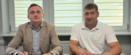 Mężczyzna w marynarce i mężczyzna w białej koszulce polo siedzą przy stole i podpisują dokumenty. Na stole leży granatowa teczka z herbem Gminy Wietrzychowice.