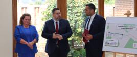 Trzy osoby, dwóch mężczyzn i kobieta stojący przy tablicy z napisem " Przebudowa wraz z rozbudową drogi w Moskorzynie". 