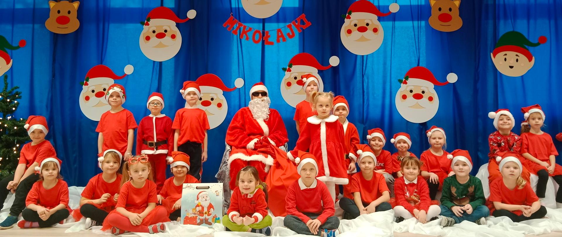 Dzieci ubrane w czerwone stroje i w czapkach mikołajkowych z Mikołajem na tle dekoracji z napisem Mikołajki