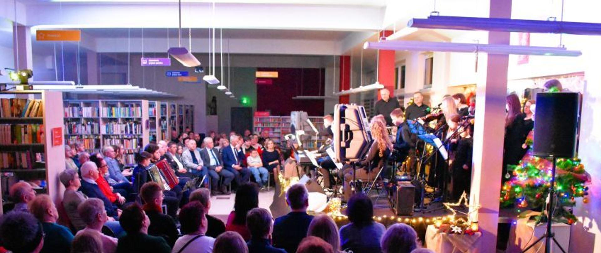 Zespół muzyczny „Owczarnia” występuje na scenie podczas Koncertu Noworocznego w bibliotece w Oleśnie. Muzycy grają i śpiewają, a zgromadzona publiczność siedzi na widowni i słucha koncertu.