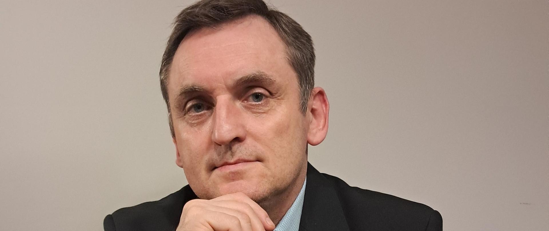 Zdjęcie przedstawia popiersie mężczyzny w wieku około 55 lat siedzącego za biurkiem. Prawą rękę ma wspartą o podbródek a lewą położoną na blacie biurka