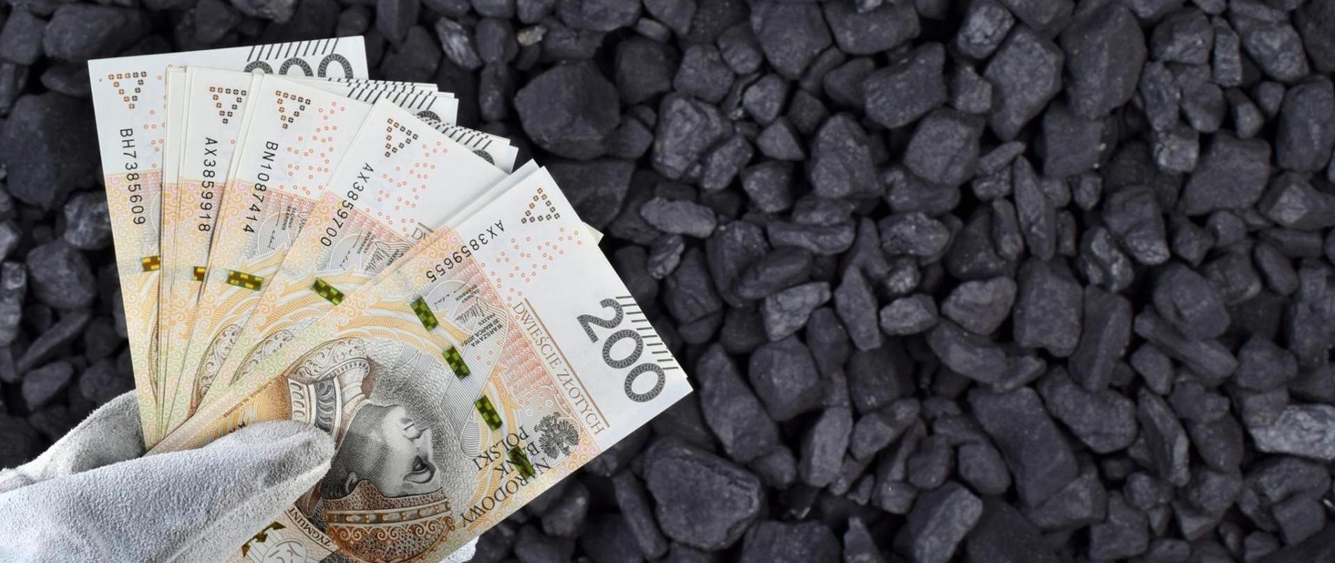 Zdjęcie dłoni w rękawicach trzymającej banknoty dwustuzłotowe na tle bryłek węgla