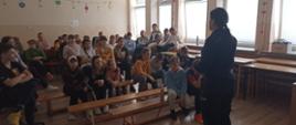 Uczniowie siedzą na ławkach i słuchają prelekcji wygłaszanej przez policjantkę 