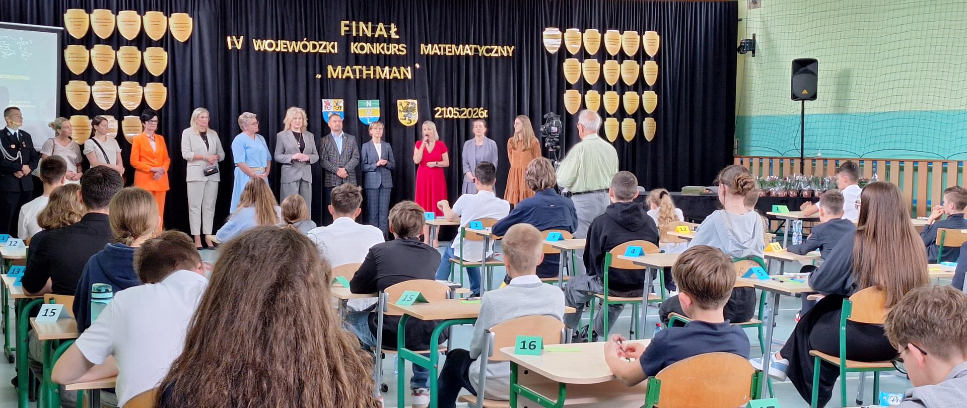 Na górze zdjęcia żółta ściana z oknami. Poniżej na ścianie granatowy materiał z napisami z liter w żółtych kolorach. Na tle materiału sylwetki osób ustawione w rzędzie. Poniżej ławki w kolorze jasnobrązowym ustawione obok siebie, przy każdej ławce sylwetki osób siedzące tyłem. Na każdej ławce kartka z numerkiem w kolorze czarnym. Z prawej strony fragment jasnej ściany w kolorze żółtym, na tle której widać siatkę.