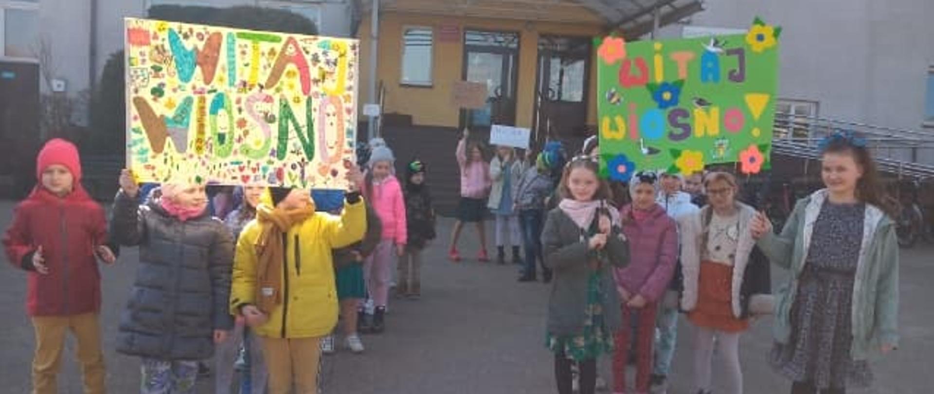 Dzieci przed budynkiem szkoły trzymają nad sobą kolorowe napisy Witaj wiosno