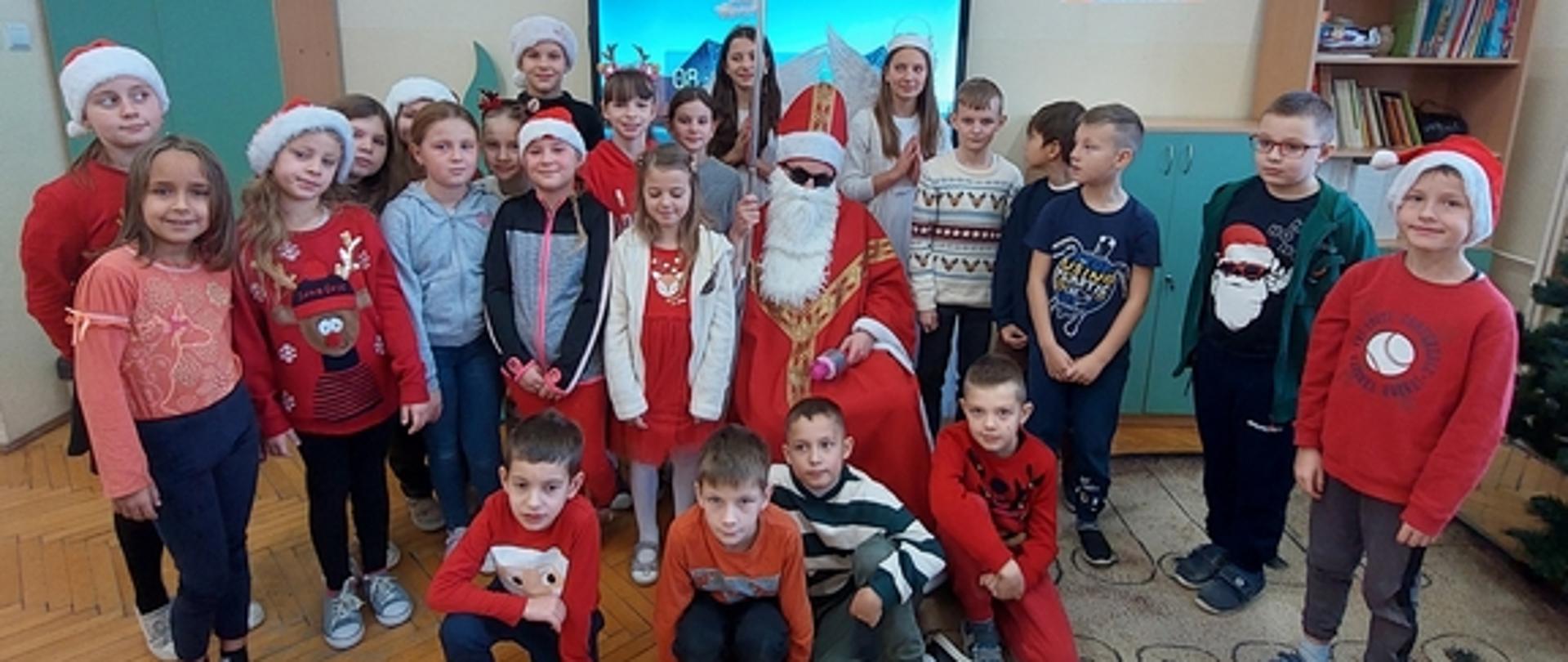 Grupka dzieci pozuje do zdjęcia w sali lekcyjnej z osobą przebraną za świętego Mikołaja i dwiema uczennicami przebranymi za aniołki. Niektóre dzieci ubrane są na kolor czerwony i mają założone na głowy czerwone czapki.
