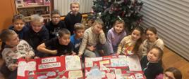 Uczniowie siedzą na podłodze w klasie szkolnej i trzymają w rękach 2 wykonane przez siebie plakaty przestawiające polskie przysłowia, za nimi choinka świąteczna