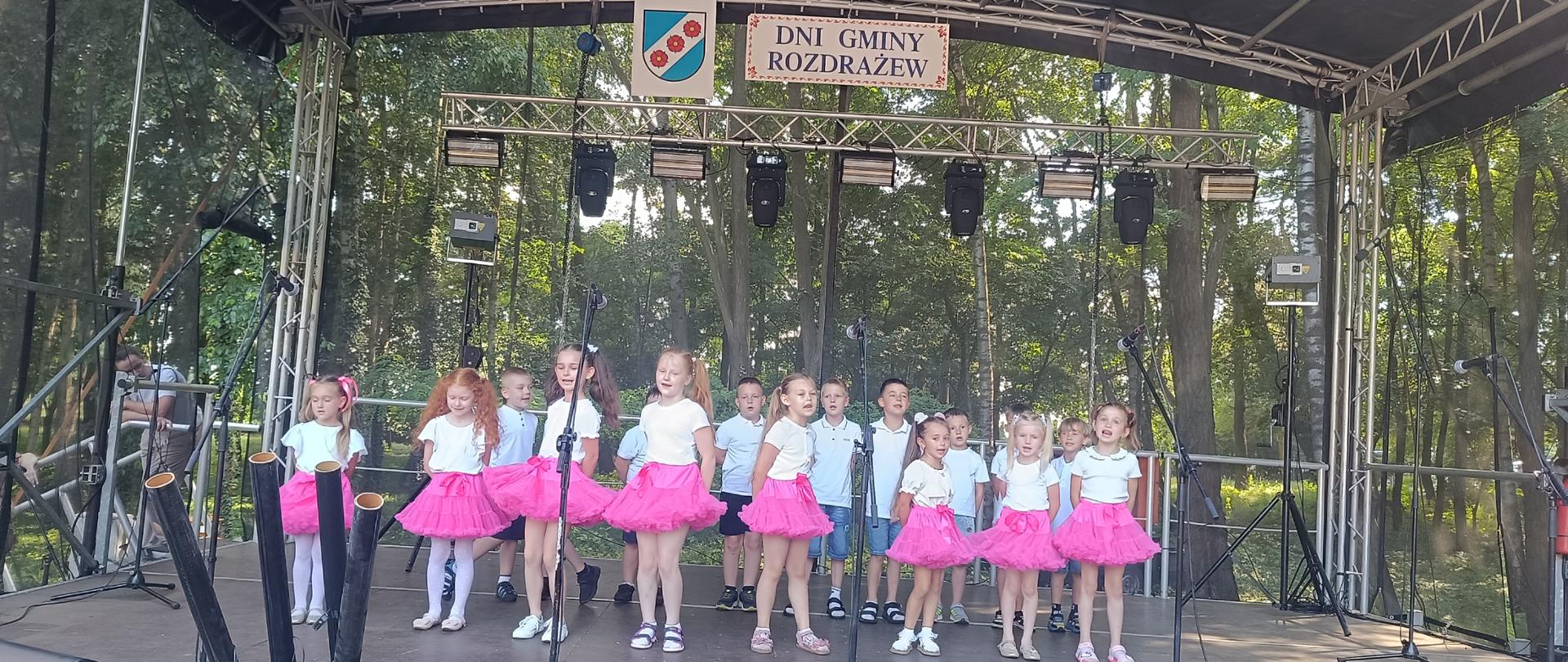 Zdjęcie przedstawia dużą scenę z zadaszeniem, sprzętem nagłaśniającym i oświetleniowym, do góry napis Dni Gminy Rozdrażew i herb Rozdrażewa. Na scenie stoją dzieci w wieku przedszkolnym w dwóch rzędach. Z przodu dziewczynki w różowych spódniczkach i białych bluzeczkach, z tyłu chłopcy w krótkich spodenkach i białych bluzkach. 