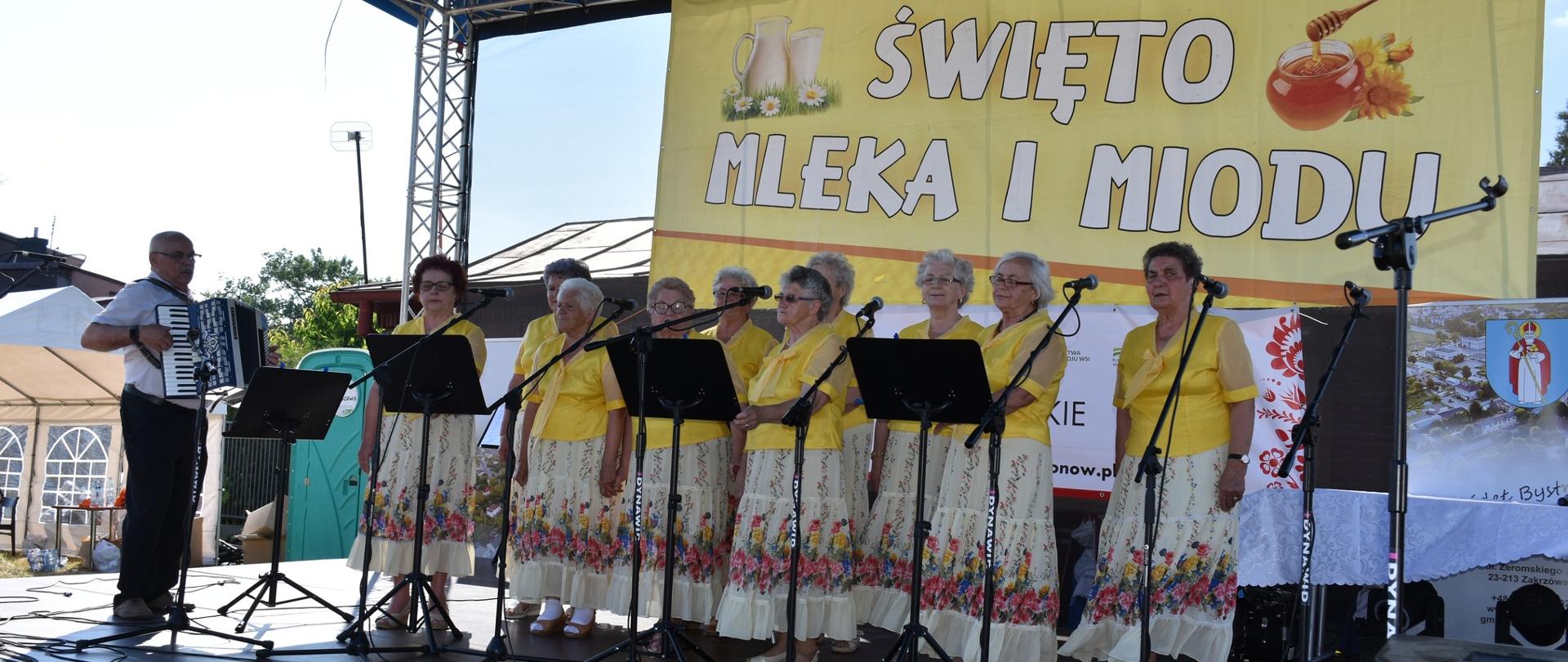 Występ zespołu ludowego podczas Święta mleka i miodu w Zakrzówku. Na zdjęciu 10 śpiewających pań i jeden pan grający na akordeonie. Panie ubrane w żółte koszulki i jasne spódnice w kwiaty.