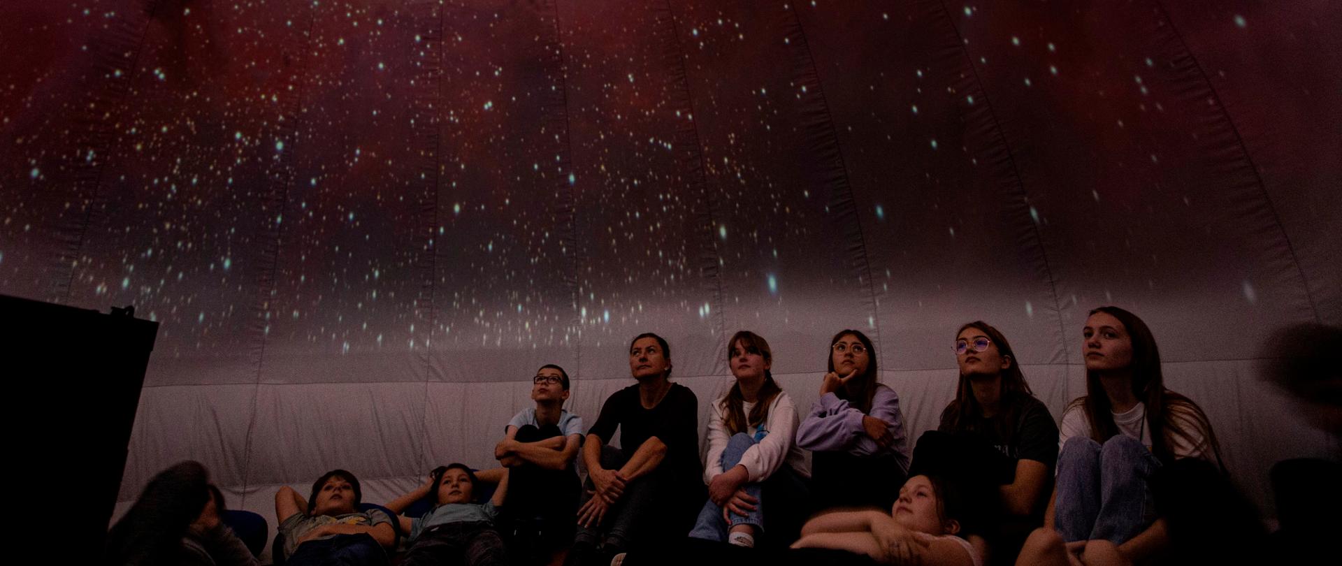 Grupa młodzieży wraz z opiekunem siedzą pod kopułą namiotu na której wyświetlany jest gwiazdozbiór.