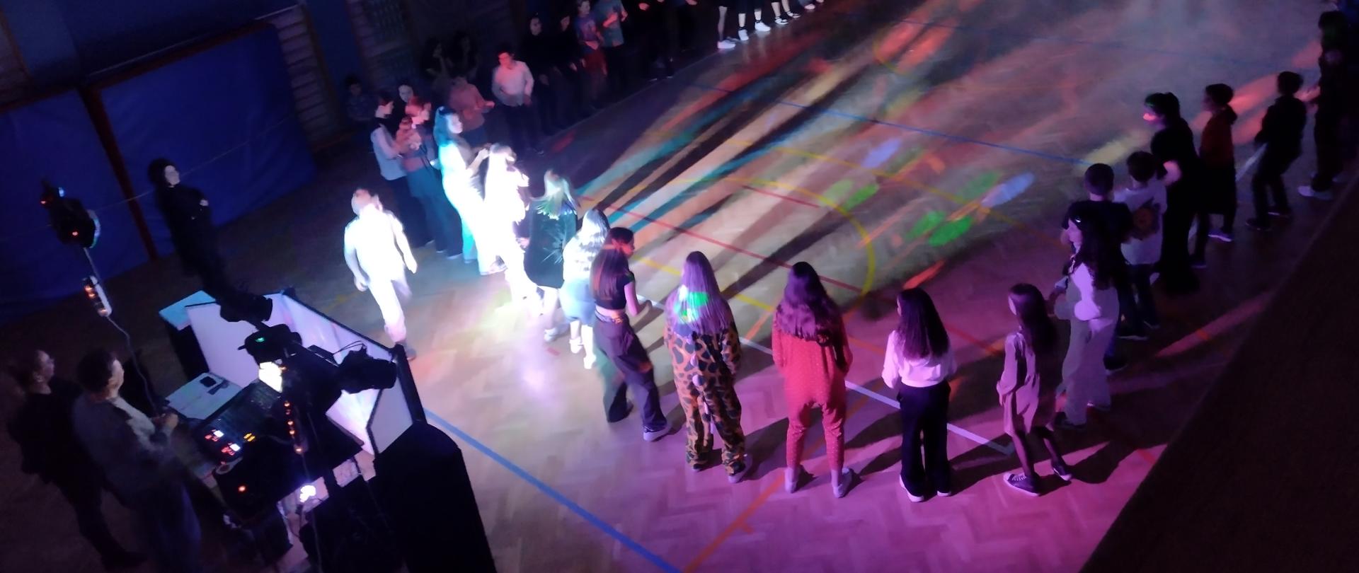 Zdjęcie przedstawia uczniów stojących w dużym kole na sali gimnastycznej i bawiących się w rytm muzyki. Z lewej strony DJ puszcza muzykę. Ciemne tło oświetlają reflektory kolorowych świateł.