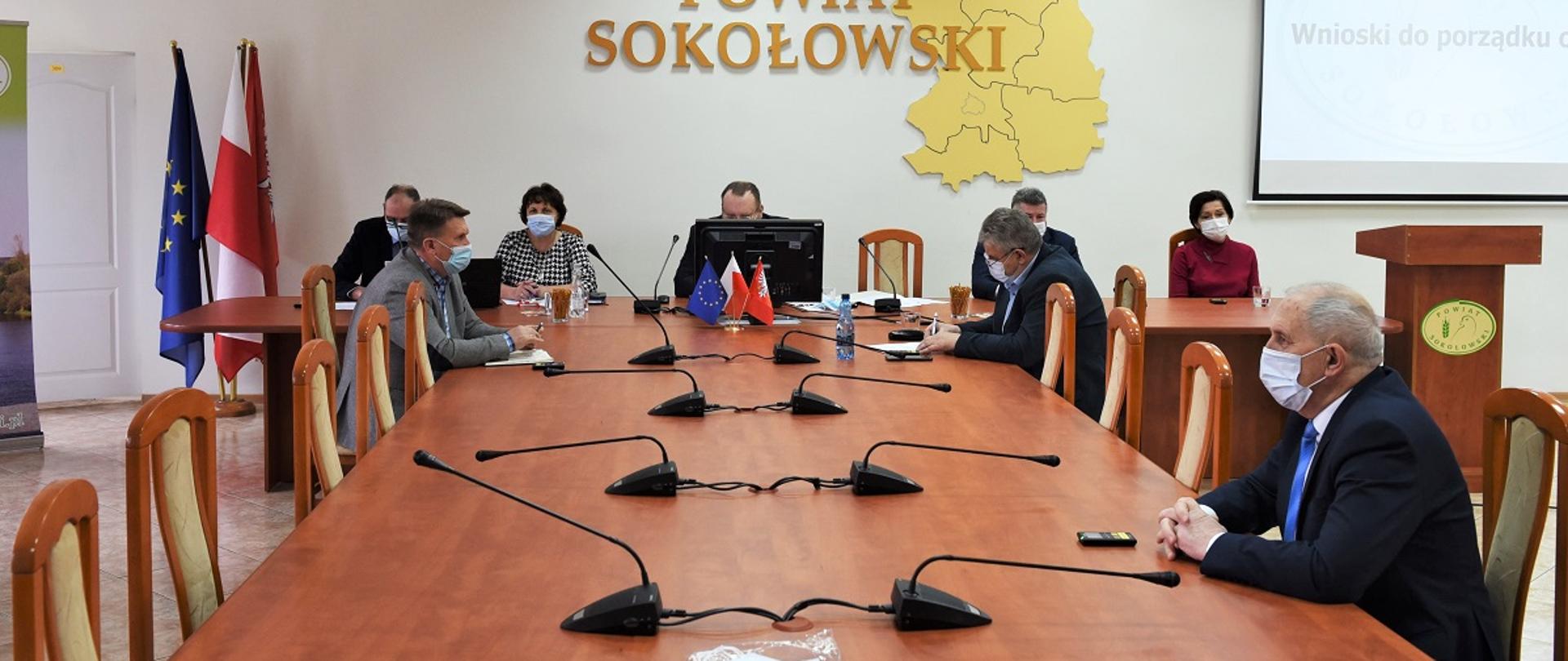 Obrady 34 sesji Rady Powiatu Sokołowskiego przeprowadzonej w hybrydowym trybie obradowania ze względu na epidemię COVID - 19. Członkowie Zarządu oraz Radni Rady Powiatu Sokołowskiego obradują przy stole obrad. W tle na ścianie złoty napis Powiat Sokołowski, krzyż katolicki oraz orzeł z godła Polski w kolorze złotym. Z boku stołu ustawione flagi Polski oraz UE