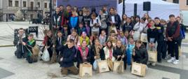 Duża grupa uczniów pozuje do wspólnej fotografii na miejskim rynku, trzymając w rękach sadzonki drzew oraz ekologiczne torby z napisem „#ekoPowiat”. W tle widać telebim z ciekawostkami na temat ekologii oraz zabytkowe kamienice, co podkreśla edukacyjny i prospołeczny charakter tego wydarzenia.