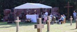 Zdjęcie wykonano z dalszej perspektywy. Widzimy polowy ołtarz przy którym ks. bp. senior odprawia mszę świętą. Na pierwszym planie na trawie rozsiane betonowe krzyże, dalej przyozdobiony kwiatami ołtarz, a w tle ściana pomnika, symbolizującego miejsce straceń, przy którym wartę pełnią żołnierze oraz harcerze.
