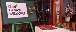 Zdjęcie przedstawia tablicę lekcyjną z napisem Dzień Edukacji Narodowej. Po prawej stronie stoi stół, na którym ułożone są dyplomy oraz róże dla nauczycieli