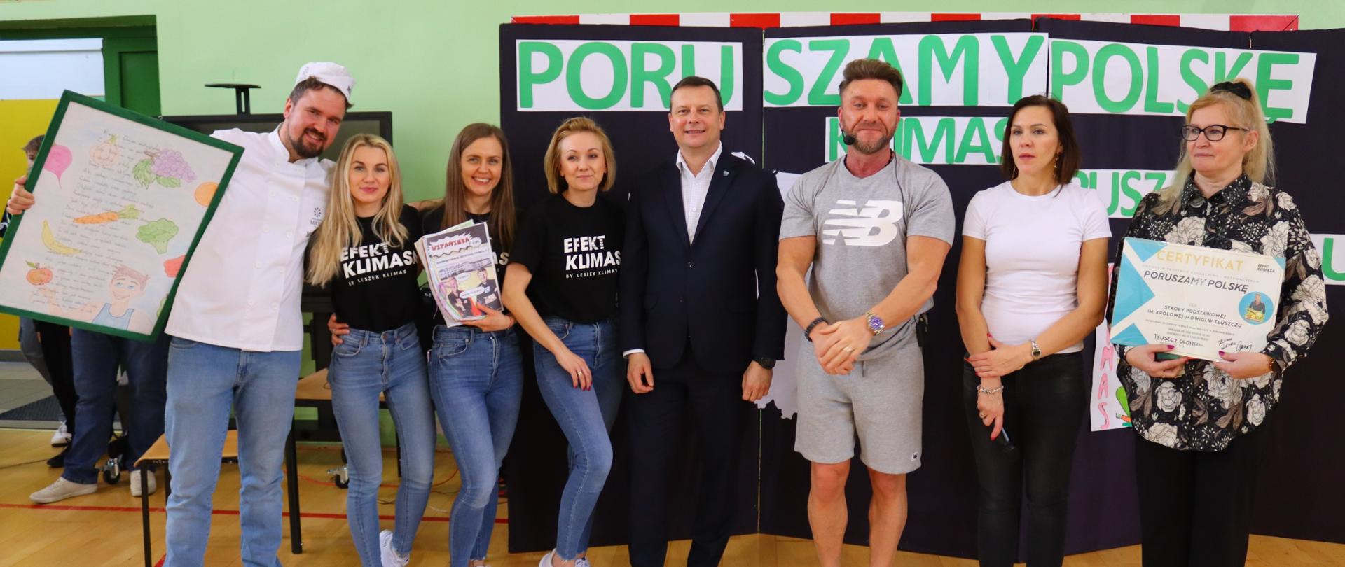 Ośmioro ludzi stojących obok siebie. W środku mężczyzna w garniturze. Po ich prawej stronie mężczyzna z białym rysunkiem w ramie