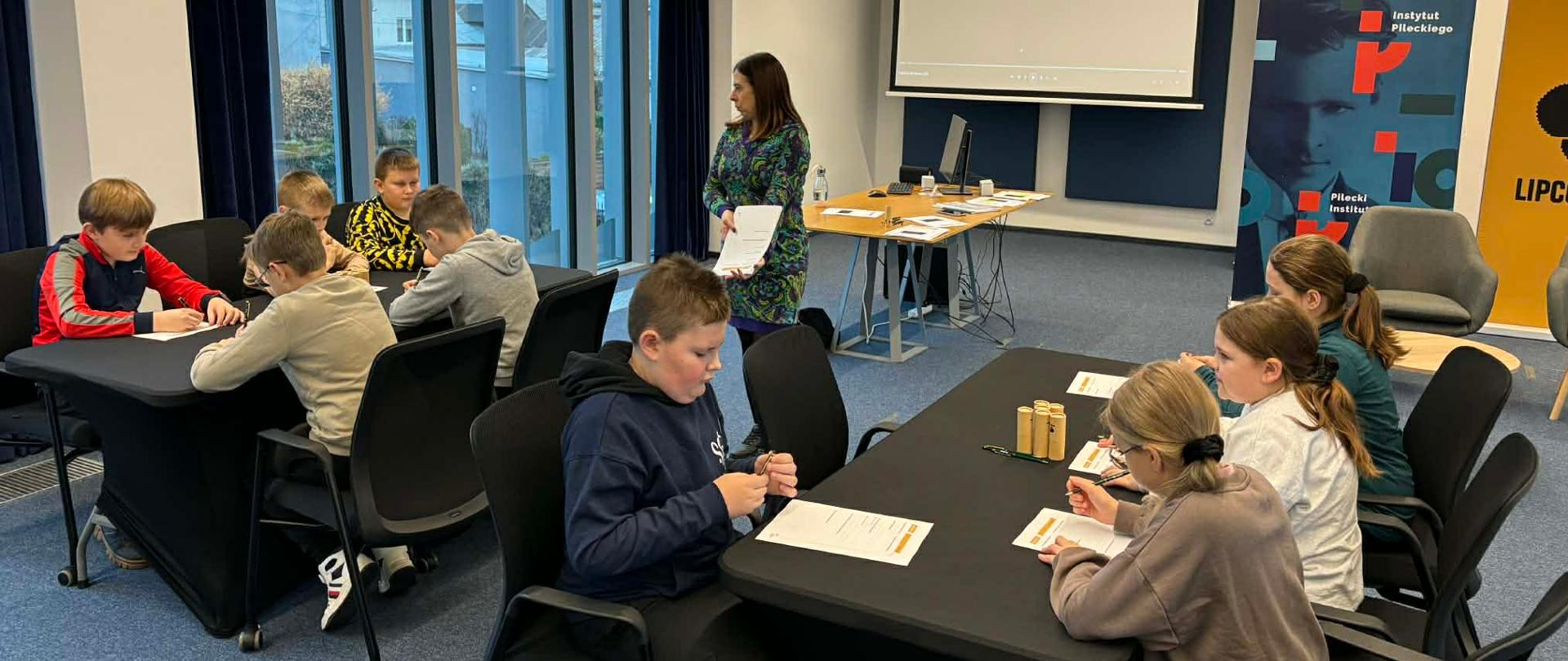 Uczniowie klasy czwartej i piątej znajdują się w sali konferencyjnej Instytutu Pileckiego w Augustowie. Dzieci siedzą na krzesłach przy dwóch stolikach. Pomiędzy nimi znajduje się kobieta. Pani trzyma w dłoni plik kartek.