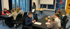 Uczniowie klasy czwartej i piątej znajdują się w sali konferencyjnej Instytutu Pileckiego w Augustowie. Dzieci siedzą na krzesłach przy dwóch stolikach. Pomiędzy nimi znajduje się kobieta. Pani trzyma w dłoni plik kartek.
