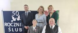 Państwo Lucyna i Krzysztof na tle granatowej tablicy z napisem wraz z Wójtem, Z-cą Wójta i Z-cą Kierownika USC