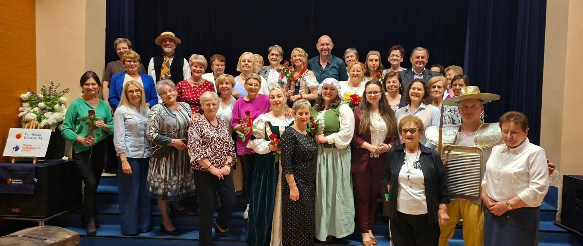 Zdjęcie przedstawia mieszkańców Gminy grodzisko Dolne ubranych odświętnie, stojących na scenie Ośrodka Kultury w Grodzisku Dolnym, uśmiechnięci