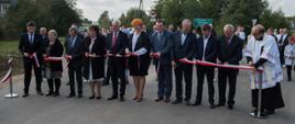 Moment przecięcia wstęgi w kolorze biało - czerwonym rozpiętej pomiędzy dwoma słupkami ustawionymi na wyremontowanej drodze. Wzdłuż wstęgi ustawieni uczestnicy otwarcia i poświęcenia i każdy przecina kawałek wstęgi. W tle mieszkańcy okolicznych miejscowości oraz wyremontowana droga.