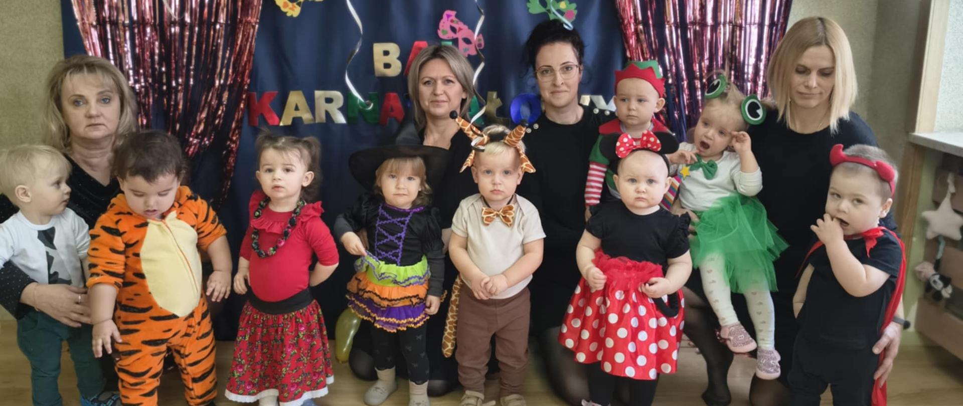 Na zdjęciu są dzieci i panie z grupy Pszczółki. Dzieci są przebrane w przeróżne stroje . W tle widnieje dekoracja w stylu karnawałowym.