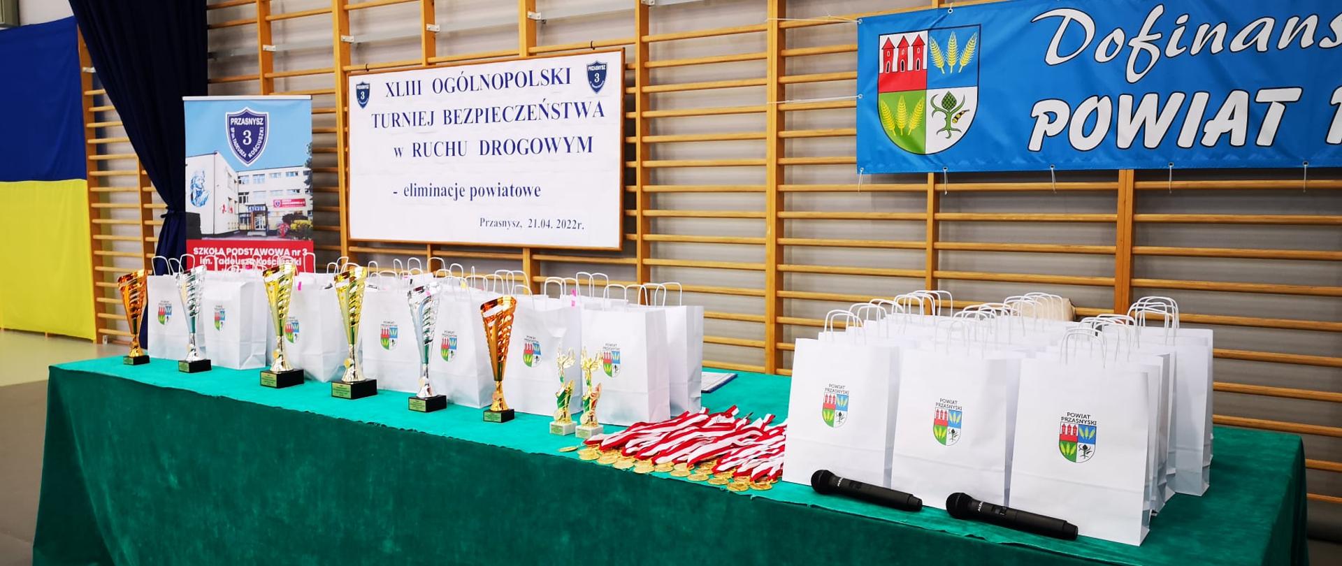 Zdjęcie przedstawia puchary, medale i nagrody czekały na uczestników powiatowych eliminacji Ogólnopolskiego Turnieju Bezpieczeństwa w Ruchu Drogowym, rozłożone na stole okrytym ciemno zielonym materiałem.