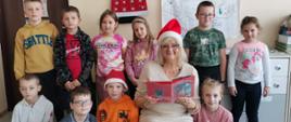 Pani bibliotekarka siedzi na krześle w czapce Mikołaja. Trzyma w rękach książeczkę „Bajkowe święta”. Wokół stoją uczniowie kl. IIa.