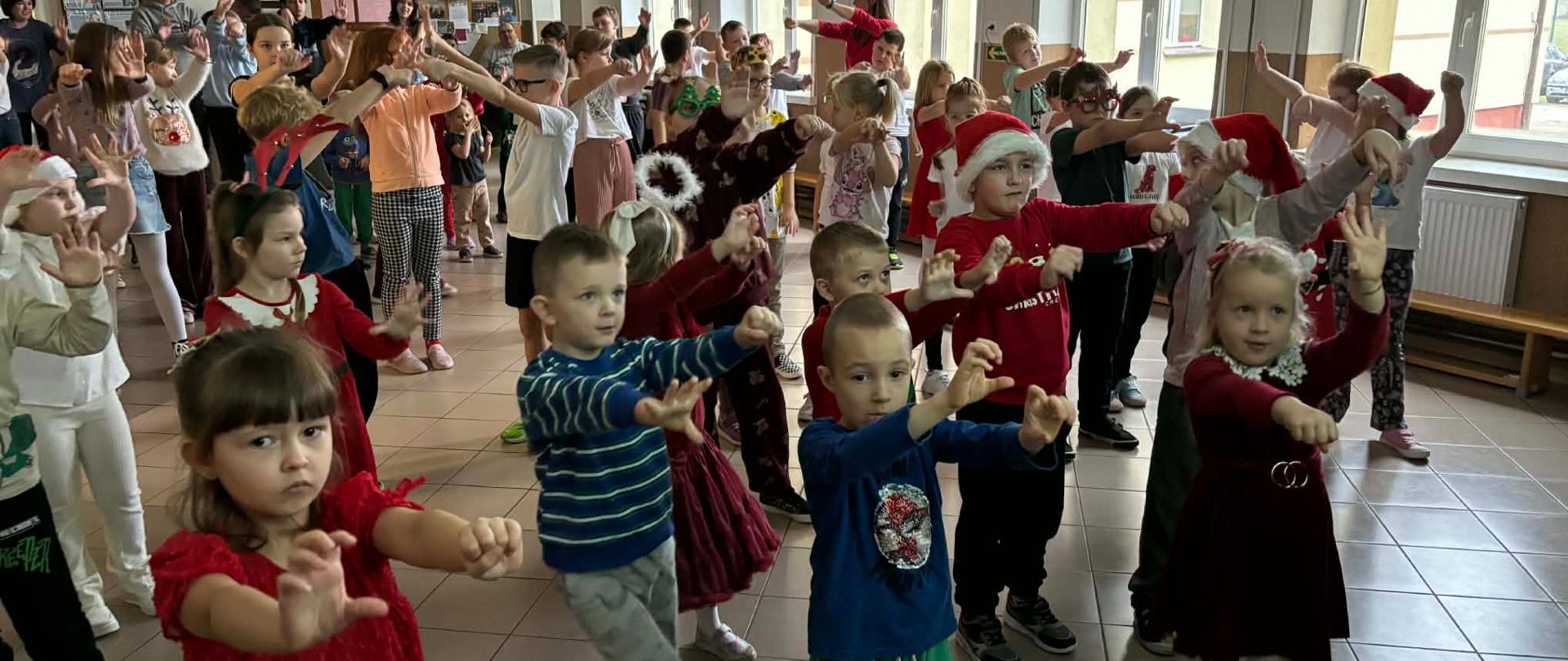 Dzieci z oddziałów przedszkolnych oraz klas młodszych wspólnie wykonują ćwiczenia gimnastyczne na korytarzu szkolnym.
