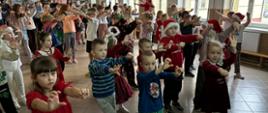 Dzieci z oddziałów przedszkolnych oraz klas młodszych wspólnie wykonują ćwiczenia gimnastyczne na korytarzu szkolnym.