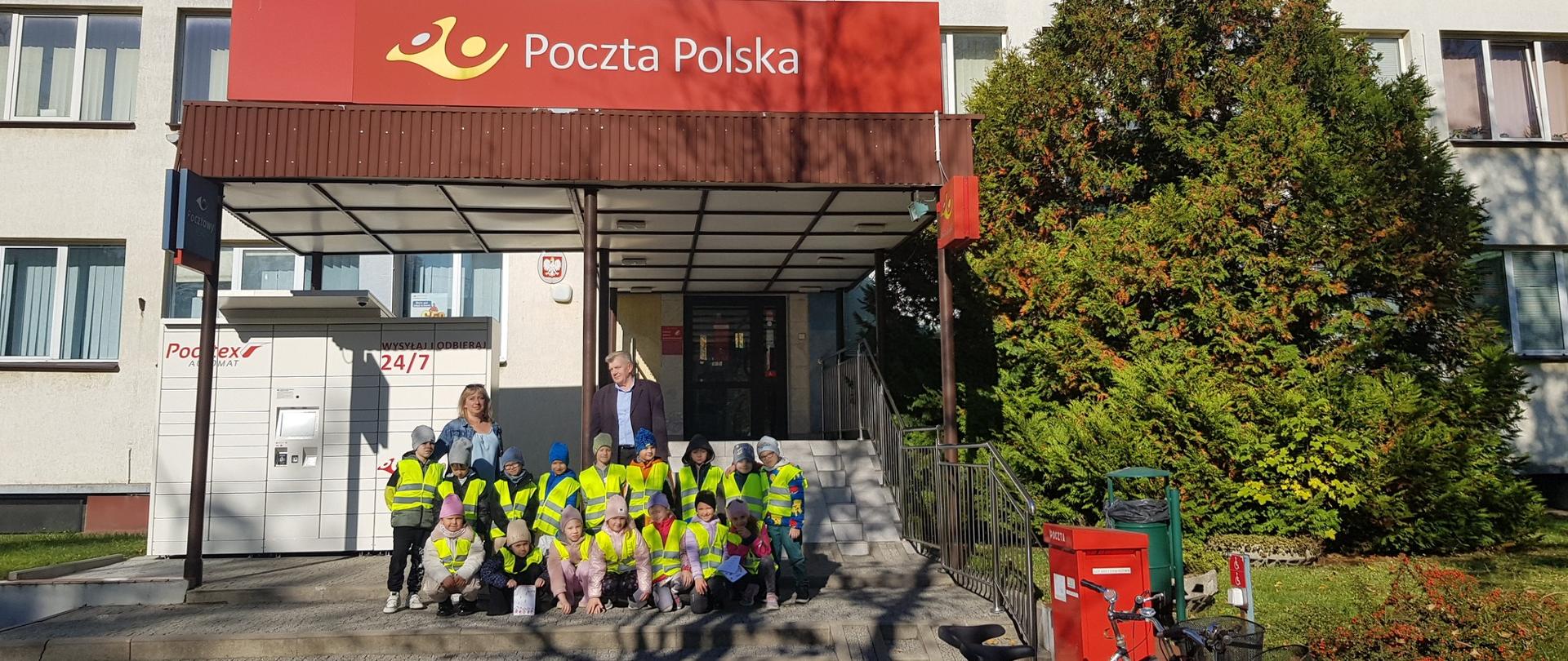 Grupa dzieci w odblaskowych koszulkach, 1 kobieta i 1 mężczyzna przed Pocztą Polską 