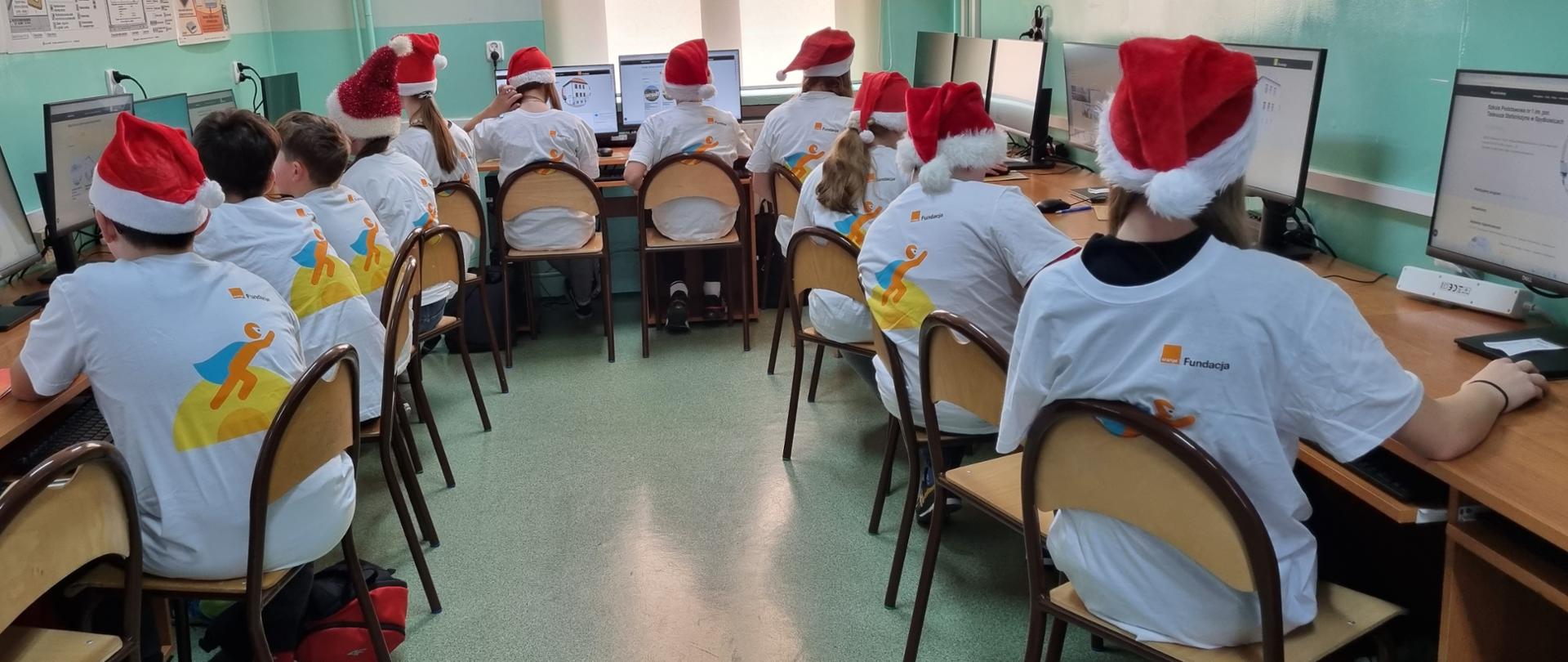 Uczniowie odwróceni tyłem do obiektywu w białych koszulkach z logo i nadrukiem #Superkoderzy, na głowie czerwone czapki Mikołaja wpatrzeni w monitor komputerów na których realizują zadania projektowe.
