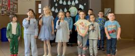 Na scenie znajdują uczniowie Oddziału Przedszkolnego A. Większość ubrana jest na niebiesko. Niektóre dzieci trzymają w dłoniach mikrofony.