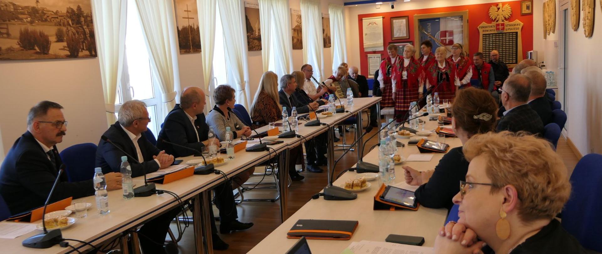 Radni Powiatu Lipskiego siedzą przy stole i wsłuchują się w występ Koła Gospodyń Wiejskich Tymienianki