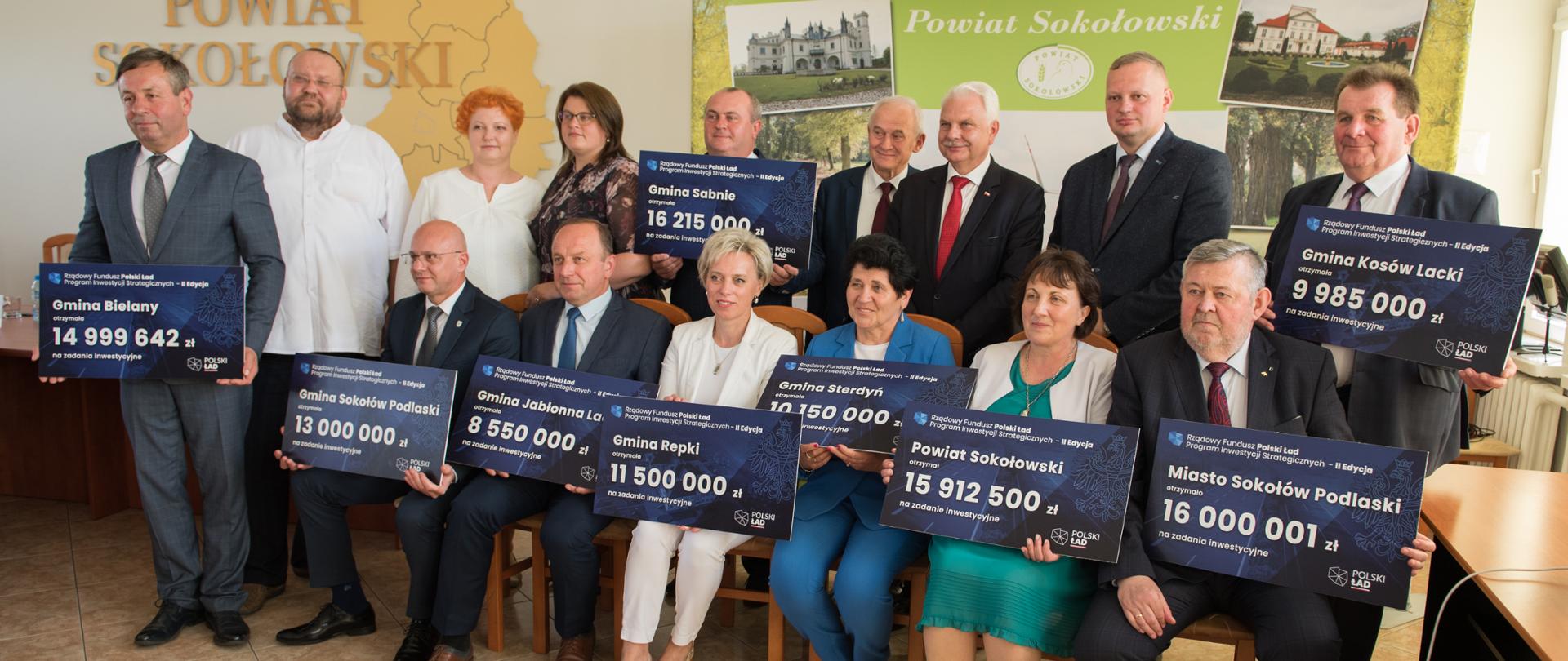 Zdjęcie grupowe wszystkich reprezentantów jednostek samorządu terytorialnego z terenu powiatu sokołowskiego wraz z parlamentarzystami oraz przedstawicielem Mazowieckiego Urzędu Wojewódzkiego. W tle widoczna ścianka promocyjna oraz napis Powiat Sokołowski wpleciony w przestrzenną mapę powiatu podzieloną na poszczególne gminy.