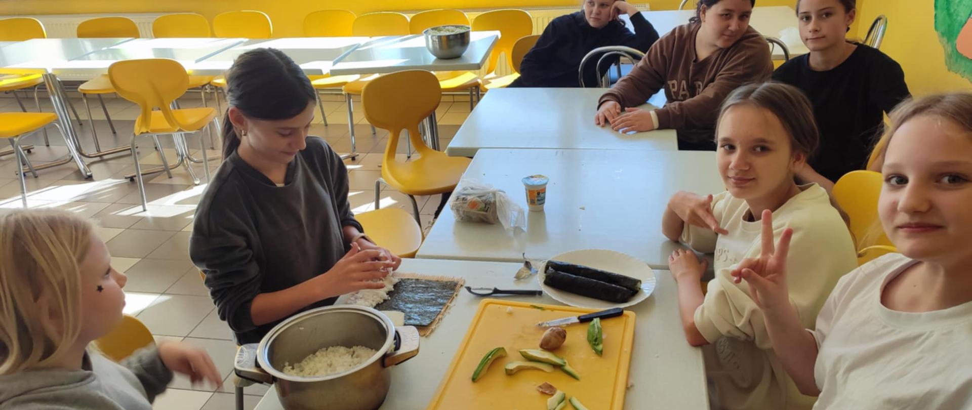 Uczennice siedzą przy stole, na którym są rolki sushi, pokrojone awokado i ogórek. Jedna uczennica zawija sushi.