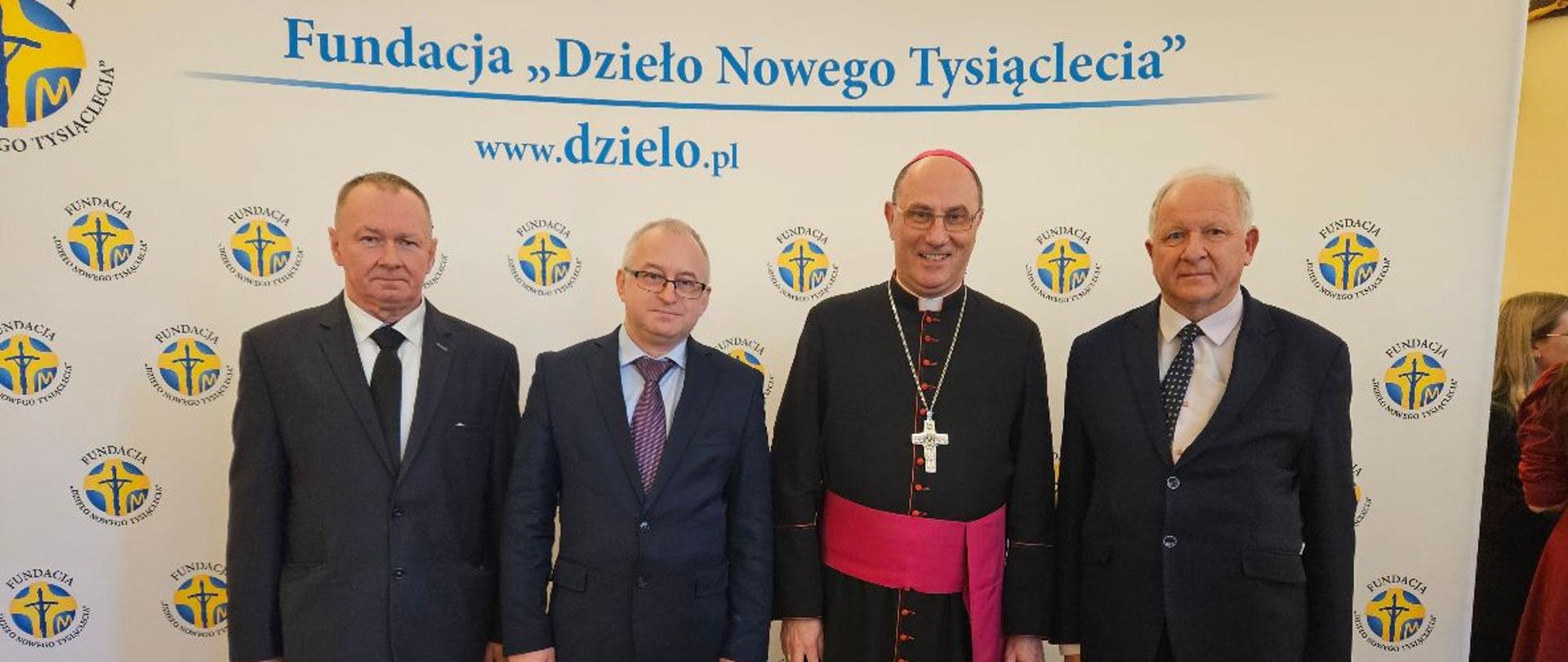 Trzech mężczyzn elegancko ubranych, stoi obok siebie uśmiechając się do zdjęcia. Po środku nich stoi kardynał Kazimierz Nycz. W tle widać baner Fundacji "Dzieło Nowego Tysiąclecia"