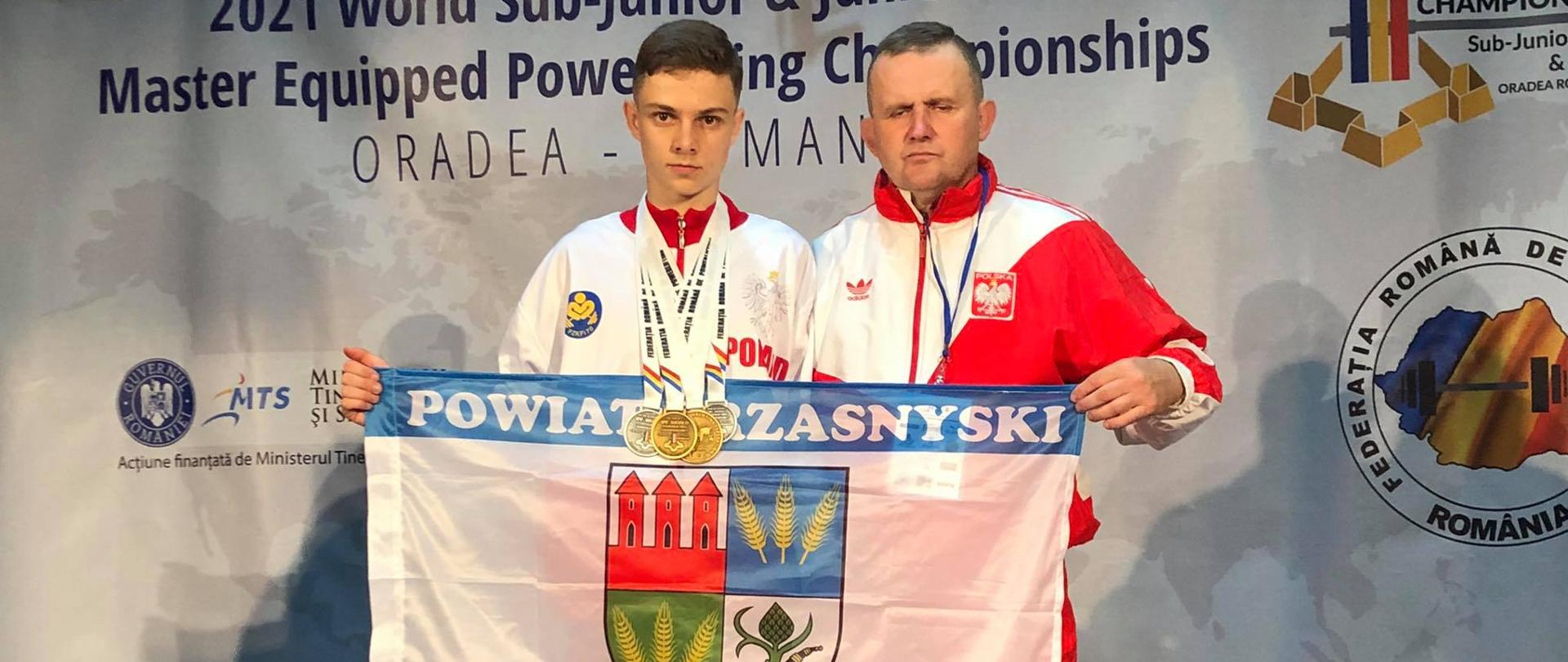 Odznaczony medalami Jakub Bońkowski (po lewej) wraz z trenerem Cezarym Pykałą (po prawej) pozują z flagą Powiatu Przasnyskiego. Zawodnik i trener trzymają biało niebieską flagę z herbem Powiatu Przasnyskiego przed sobą.