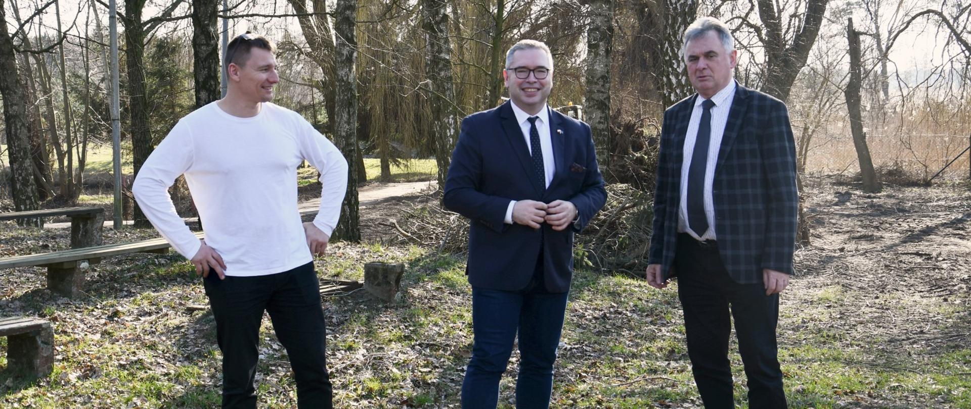 Zdjęcie przedstawiające trzech mężczyzn stojących w zalesionym terenie, na skraju modernizowanego amfiteatru przy MGOK w Margoninie.
Od lewej stoją:
Łukasz Malczewski – Dyrektor Miejsko-Gminnego Ośrodka Kultury w Margoninie, w białej bluzce z długim rękawem i ciemnych spodniach.
Jacek Bogusławski – Członek Zarządu Województwa Wielkopolskiego, w ciemnym garniturze, białej koszuli i krawacie.
Janusz Piechocki – Burmistrz Margonina, w marynarce w kratę, białej koszuli i krawacie.
Osoby są uśmiechnięte i patrzą w stronę obiektywu. Stoją na skraju wyrównanego terenu, otoczeni drzewami w stanie bezlistnym. W tle po lewej widać starsze drewniane ławki. Kontekst spotkania dotyczy modernizacji amfiteatru w ramach dofinansowania „Kulisy Kultury”.