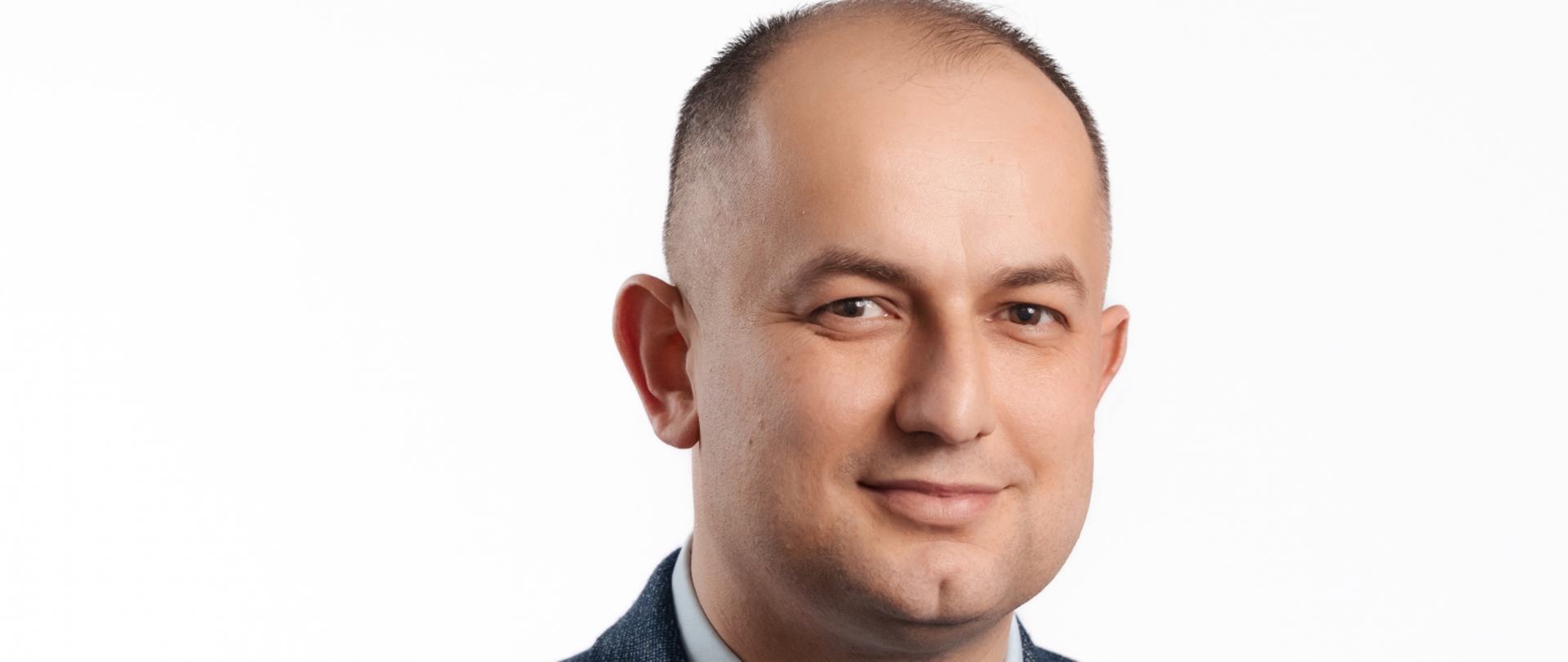Lekko łysiejący szatyn, wąskie brązowe oczy, delikatny uśmiech, charakterystyczny przecinek na brodzie. Ubrany w granatową marynarkę, jasnoniebieska koszulę, ciemnoniebieski krawat. 
