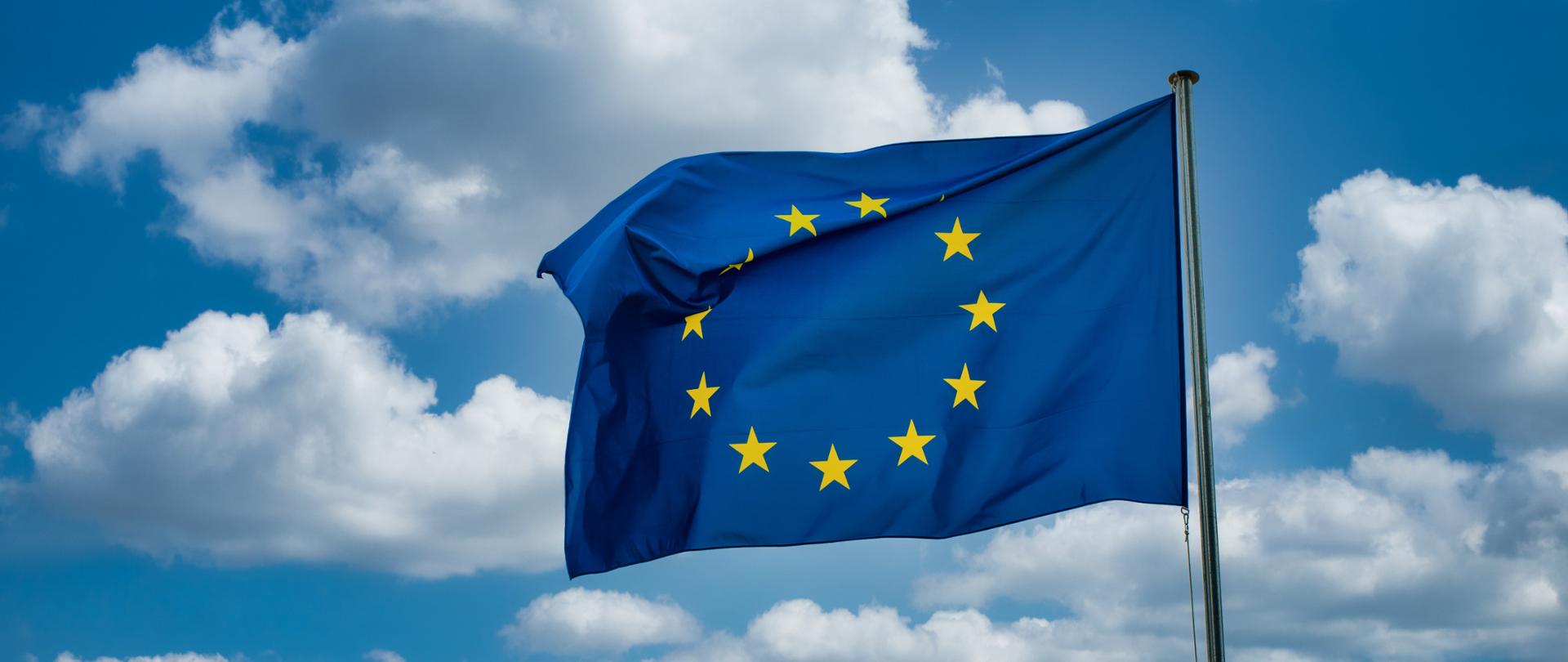 Zdjęcie przedstawia niebieską z żółtymi gwiazdkami flagę Unii Europejskiej zawieszoną na maszcie na tle zachmurzonego nieba.