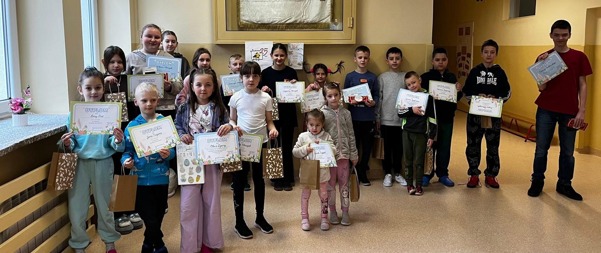 Na korytarzu pod gablotą zawierającą sztandar szkoły stoją uczniowie nagrodzenie w konkursie, trzymają w ręku dyplomy oraz ozdobne torebeczki z nagrodami