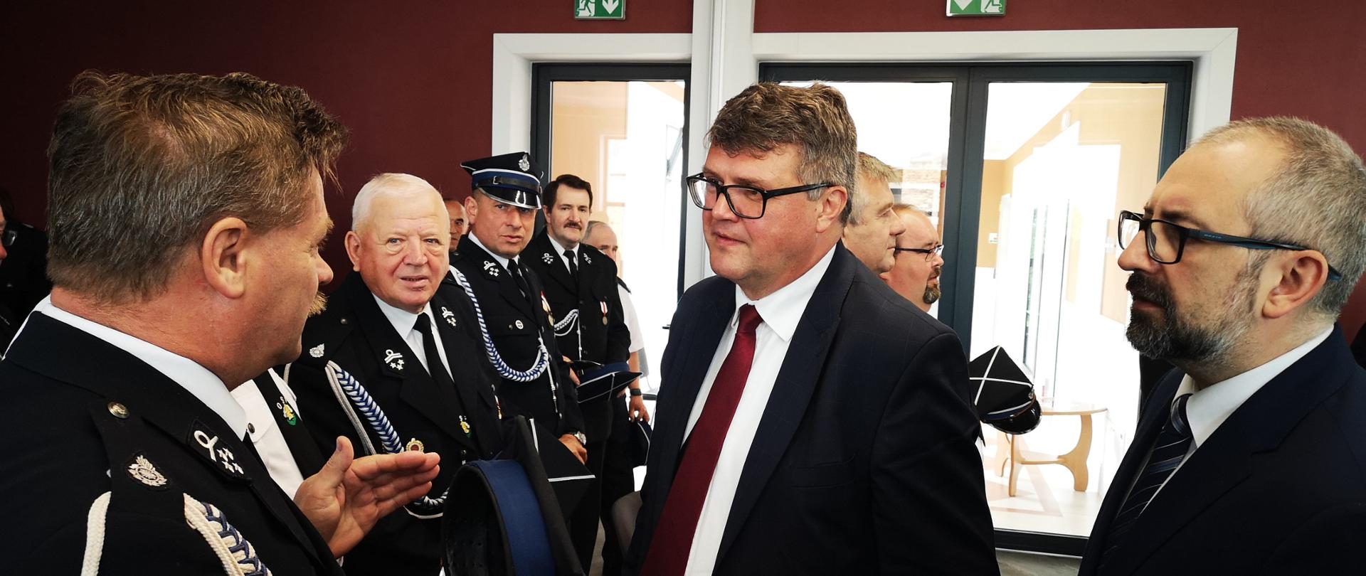 Zdjęcie przedstawia Ministra Macieja Wąsika oraz Starostę Przasnyskiego Krzysztofa Bieńkowskiego witających się ze strażakami z terenu powiatu przasnyskiego. Spotkanie miało miejsce na sali konferencyjnej w Centrum Aktywizacji Biznesu w budynku Starostwa Powiatowego w Przasnyszu.