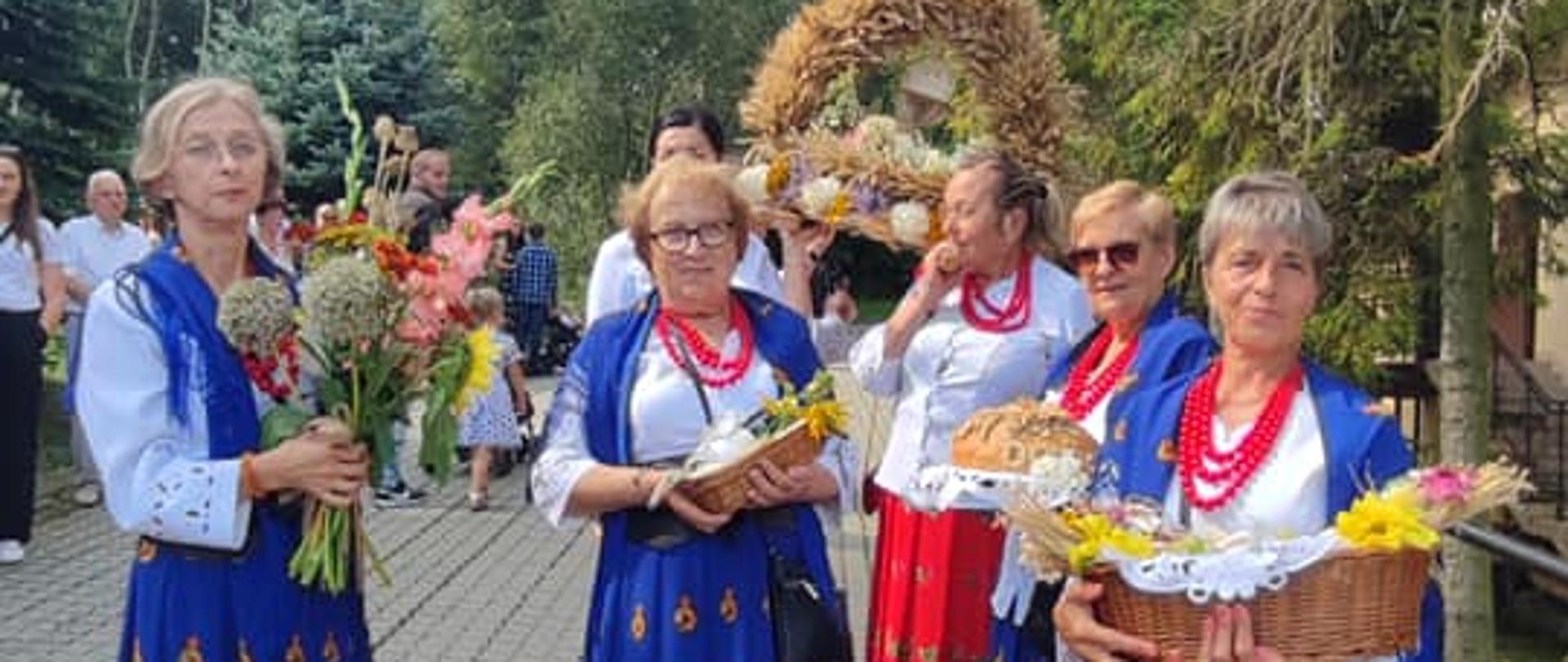Kobiety ubrane w stroje regionalne trzymające dary dożynkowe. W tle wieniec dożynkowy