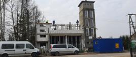 Remontowany budynek remizy OSP w Trzciance. Przed posesją niebieski kontener oraz dwa samochody.