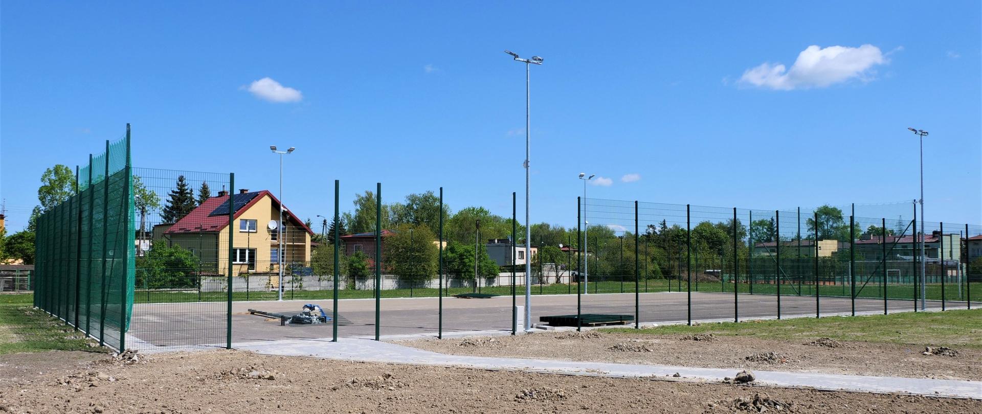 Zdjęcie przedstawia budowę boiska. Boisko ogrodzone siatką, a na powierzchni wylany jest asfalt. Boisko otoczone jest usypaną ziemią.