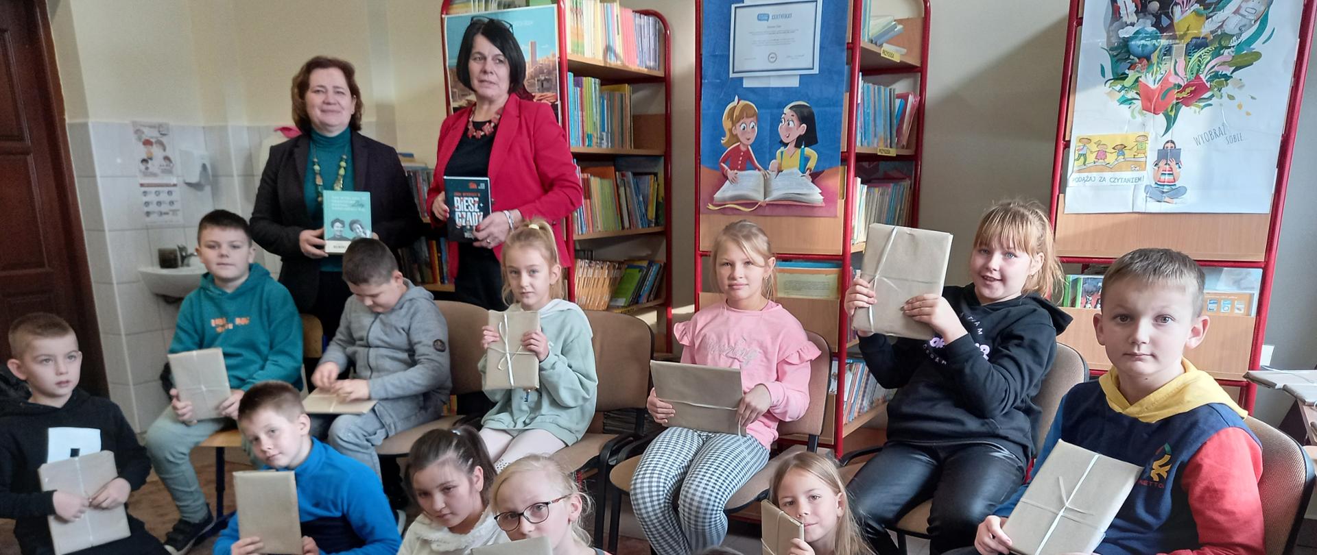 Zdjęcie grupowe kl.2b wraz z wychowawczynią i Panią dyrektor w bibliotece szkolnej. Wszyscy trzymają w rękach zapakowaną w szary papier "książkę – niespodziankę".