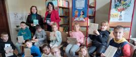 Zdjęcie grupowe kl.2b wraz z wychowawczynią i Panią dyrektor w bibliotece szkolnej. Wszyscy trzymają w rękach zapakowaną w szary papier "książkę – niespodziankę".