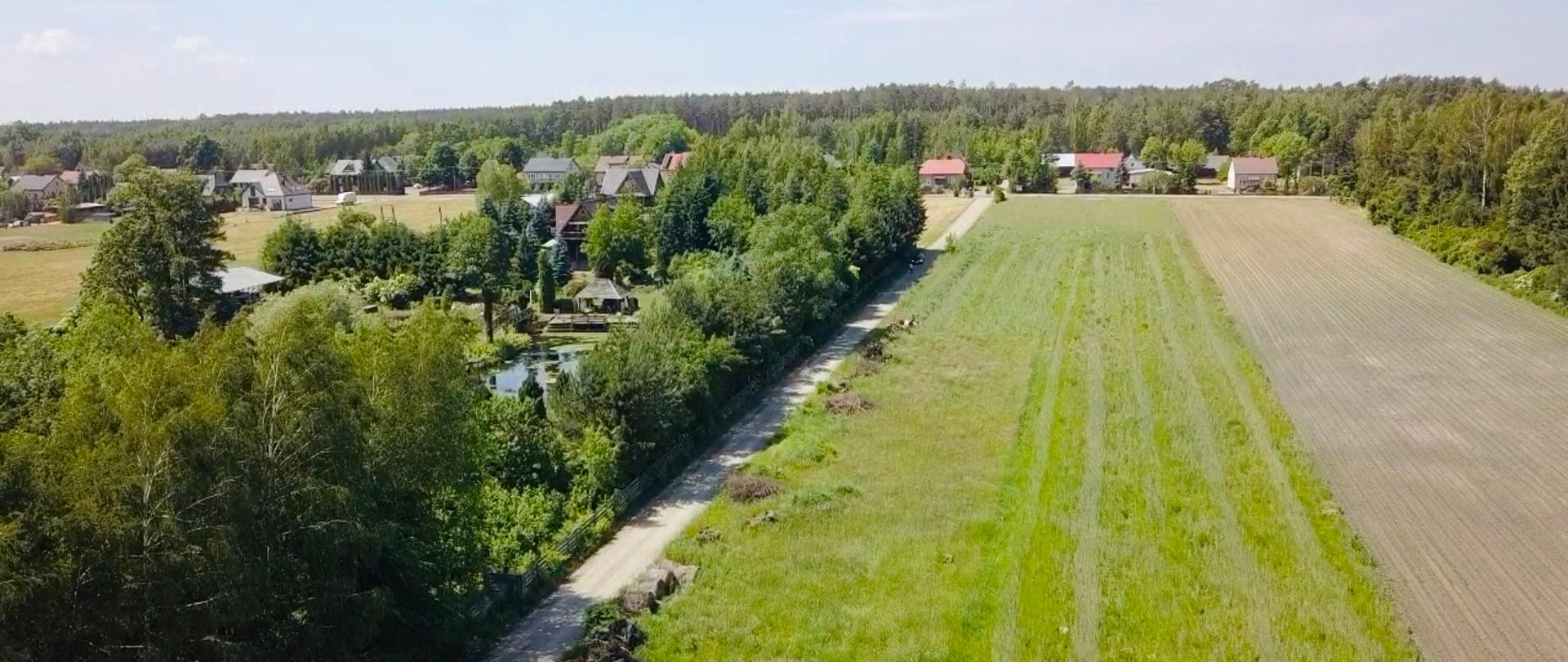 Widok z lotu ptaka, droga gruntowa, po prawej stronie łąka i pole. Po lewej stronie zadrzewienia i zabudowania. w oddali inne zabudowania oraz las.