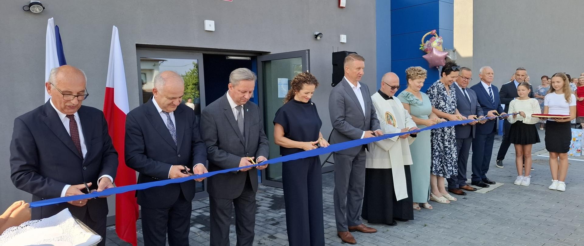 Kilkanaście dorosłych osób i dwie dziewczynki wspólnie przecinają niebieską wstęgę. Wstęga jest przytrzymywana przez osoby na skrajach rzędu. Wszyscy są elegancko ubrani, a tło stanowią szary budynek oraz polskie i unijne flagi. Na fasadzie budynku widnieje napis.